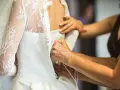 Dettaglio di abiti da sposa riutilizzabili: mani che chiudono bottoncini su un vestito bianco con pizzo e velo.