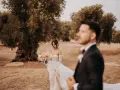 Sposarsi a maggio sotto un ulivo secolare. La sposa con un bouquet di rose pesca, lo sposo in primo piano sfocato.