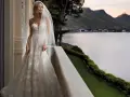 Sposa con abito da sogno, con vari tipi di pizzo che scendono a cascata, su un balcone con vista lago al tramonto.