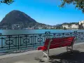 Panoramica del lago di Lugano con Monte San Salvatore sullo sfondo. Una panchina rossa invita a godersi la vista, perfetta per scoprire cosa vedere a Lugano in un giorno.