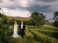 Sposi passeggiano mano nella mano tra i verdi labirinti della **villa di Maiano**, con un bouquet fiorito e uno sfondo di colline toscane.