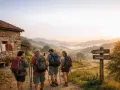 Pellegrini con zaini e bastoncini da trekking si dirigono verso un cartello che indica "Santiago Camino". La domanda "quanto dura il cammino di Santiago" risuona mentre il sole sorge su dolci colline nebbiose.