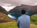 Uomo con maglione grigio ammira un paesaggio mozzafiato delle Isole Faroe, con un lago turchese e una cascata.