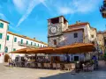 Piazza nel kotor centro storico con torre dell'orologio e tavoli all'aperto di un ristorante.