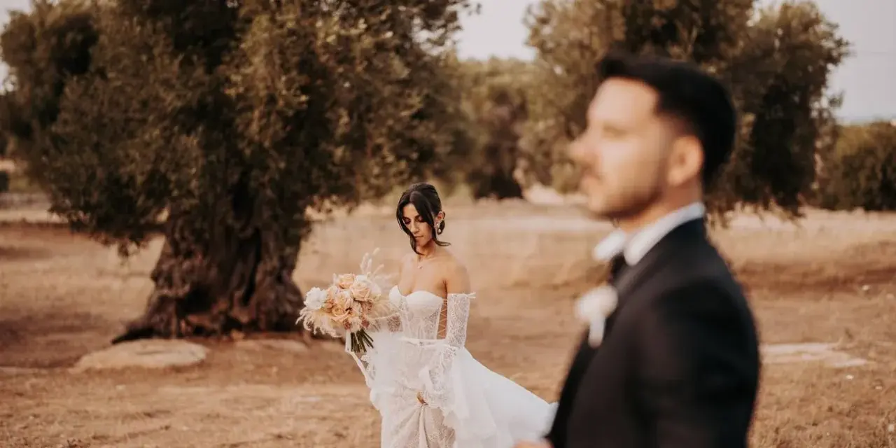 Sposarsi a maggio sotto un ulivo secolare. La sposa con un bouquet di rose pesca, lo sposo in primo piano sfocato.