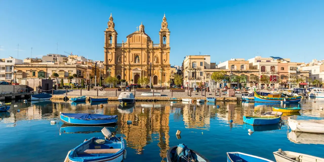Barche colorate nel porto di Marsaxlokk, Malta. Un luogo incantevole dove dormire a Malta, con la chiesa barocca che domina lo sfondo.