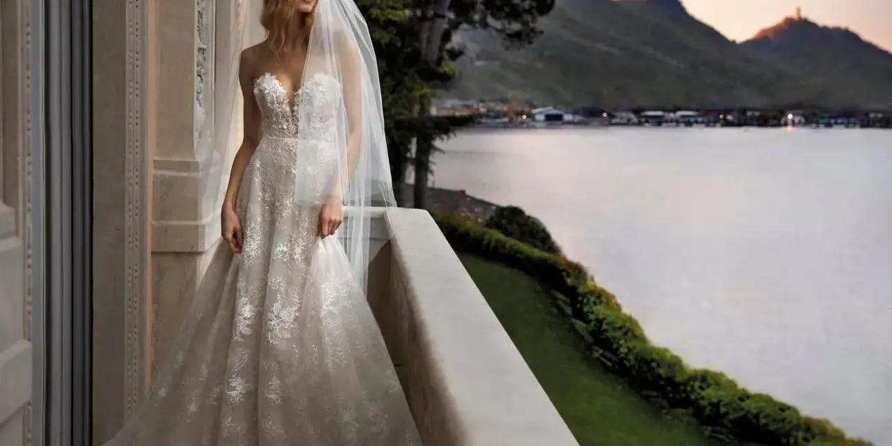 Sposa con abito da sogno, con vari tipi di pizzo che scendono a cascata, su un balcone con vista lago al tramonto.