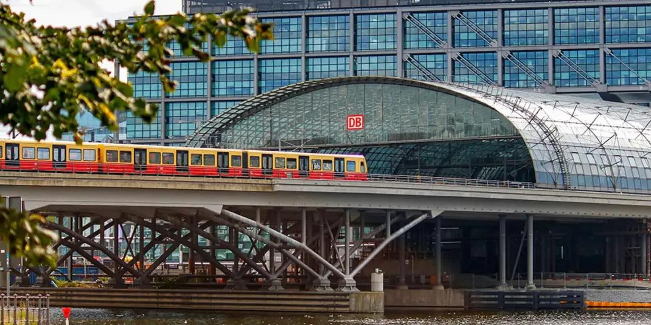 Treno giallo e rosso attraversa un ponte sopra un fiume, con la moderna stazione ferroviaria di Berlino sullo sfondo. Un ottimo modo per capire come muoversi a Berlino.