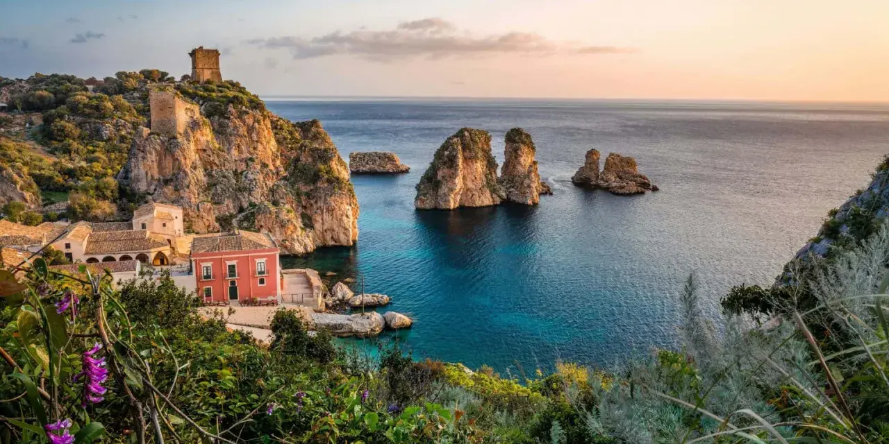 Un borgo con torri antiche e un edificio rosso si affaccia su un mare cristallino, perfetto per le mete estive in Italia.
