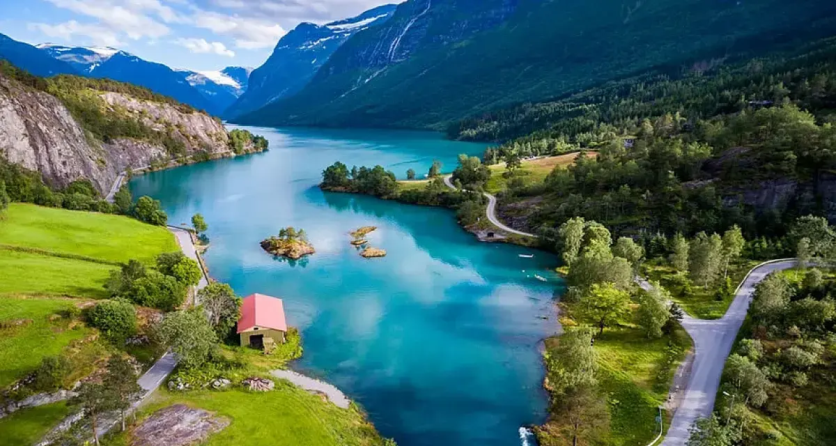 Un lago turchese serpeggia tra montagne verdi e innevate. Un piccolo edificio rosso si affaccia sull'acqua. La crociera fiordi periodo migliore è l'estate per godere di questi panorami.