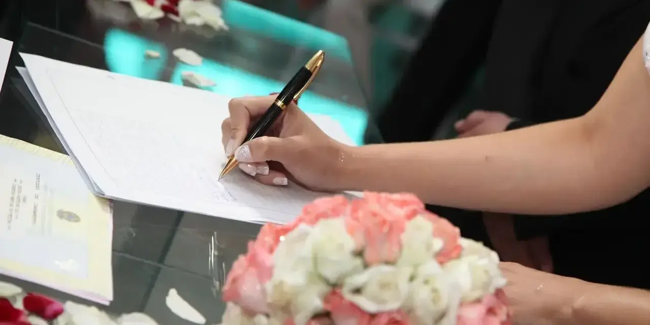 Mano di una sposa che firma un documento, forse un accordo di separazione dei beni, con un bouquet di rose in primo piano.