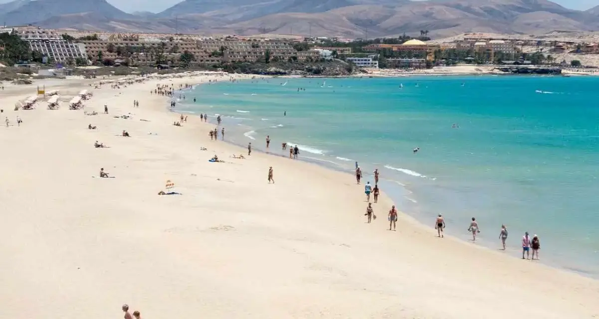 Spiaggia affollata con persone che passeggiano e si rilassano, hotel e montagne sullo sfondo. Questo è il paesaggio di Fuerteventura, dove si trova questo paradiso.