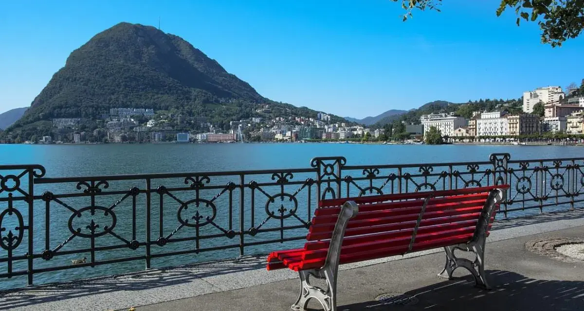 Panoramica del lago di Lugano con Monte San Salvatore sullo sfondo. Una panchina rossa invita a godersi la vista, perfetta per scoprire cosa vedere a Lugano in un giorno.