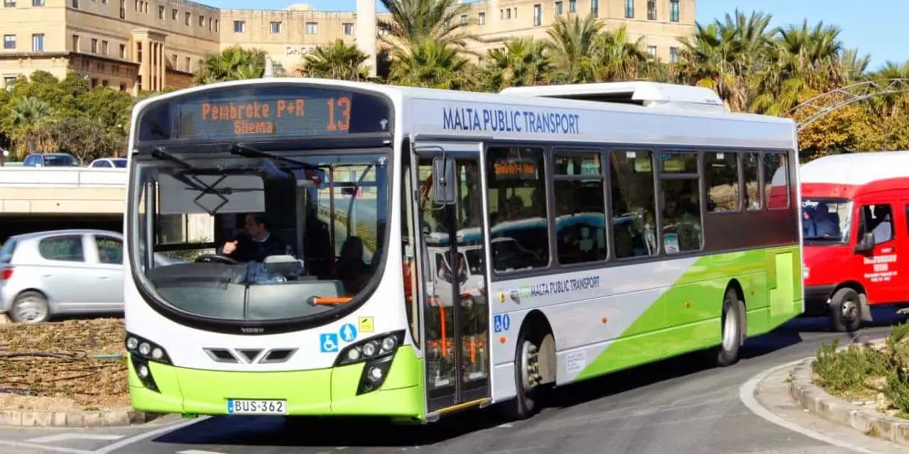 Autobus bianco e verde di Malta Public Transport, ideale per scoprire come spostarsi a Malta. Percorre una strada con palme e edifici storici.