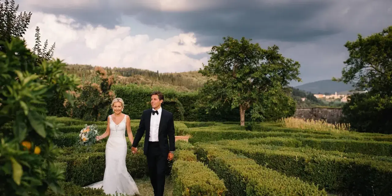 Sposi passeggiano mano nella mano tra i verdi labirinti della **villa di Maiano**, con un bouquet fiorito e uno sfondo di colline toscane.