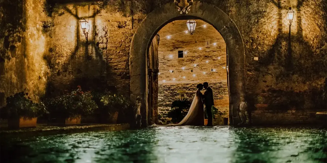 Una coppia di sposi si abbraccia sotto un arco del castello di San Basilio, illuminato da lanterne e luci soffuse. L'acqua riflette l'atmosfera magica.