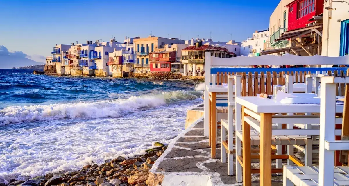 Case vacanza a Mykonos, un'idea per dove andare a vivere all'estero con pochi soldi. Case bianche sul mare, onde che si infrangono sugli scogli, tavoli di un ristorante.