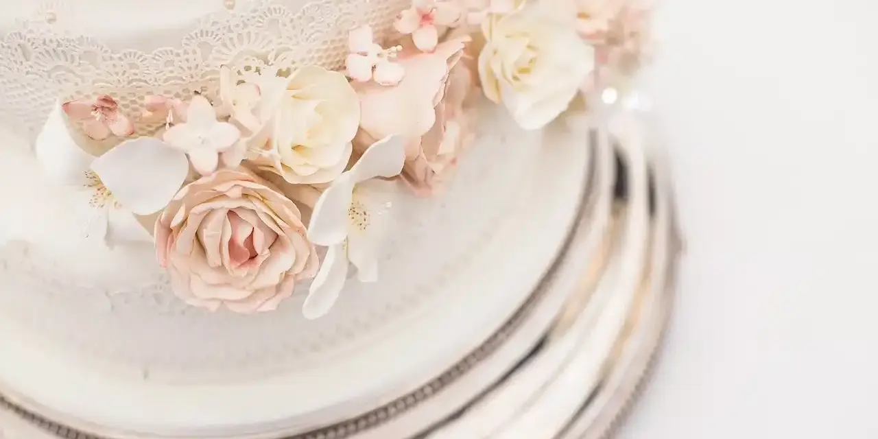 Dettaglio di una torta nuziale a più piani, decorata con pizzo e fiori di zucchero rosa e bianchi, su un elegante vassoio argentato.
