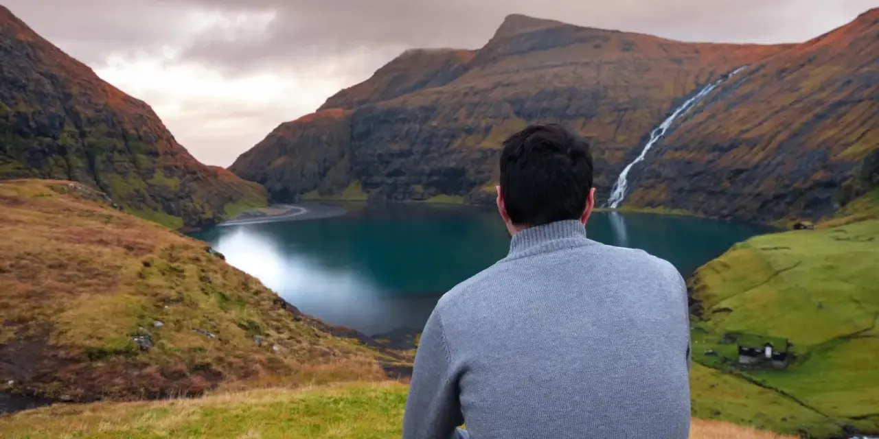 Uomo con maglione grigio ammira un paesaggio mozzafiato delle Isole Faroe, con un lago turchese e una cascata.