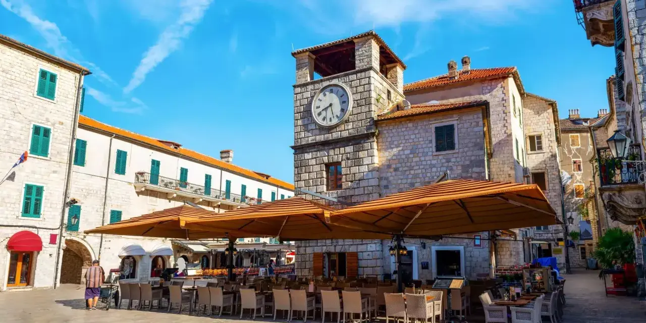 Piazza nel kotor centro storico con torre dell'orologio e tavoli all'aperto di un ristorante.