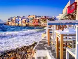 Case vacanza a Mykonos, un'idea per dove andare a vivere all'estero con pochi soldi. Case bianche sul mare, onde che si infrangono sugli scogli, tavoli di un ristorante.