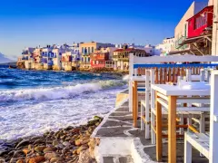Case vacanza a Mykonos, un'idea per dove andare a vivere all'estero con pochi soldi. Case bianche sul mare, onde che si infrangono sugli scogli, tavoli di un ristorante.