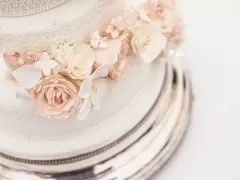 Dettaglio di una torta nuziale a più piani, decorata con pizzo e fiori di zucchero rosa e bianchi, su un elegante vassoio argentato.