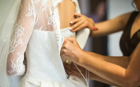 Dettaglio di abiti da sposa riutilizzabili: mani che chiudono bottoncini su un vestito bianco con pizzo e velo.