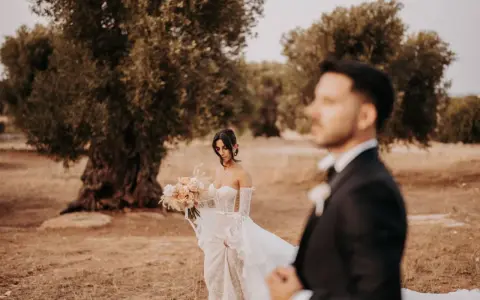 Sposarsi a maggio sotto un ulivo secolare. La sposa con un bouquet di rose pesca, lo sposo in primo piano sfocato.