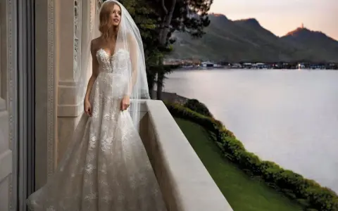 Sposa con abito da sogno, con vari tipi di pizzo che scendono a cascata, su un balcone con vista lago al tramonto.