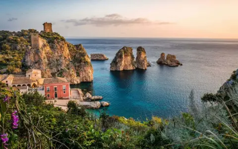 Un borgo con torri antiche e un edificio rosso si affaccia su un mare cristallino, perfetto per le mete estive in Italia.