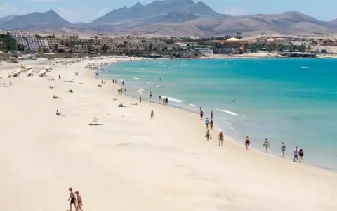Spiaggia affollata con persone che passeggiano e si rilassano, hotel e montagne sullo sfondo. Questo è il paesaggio di Fuerteventura, dove si trova questo paradiso.