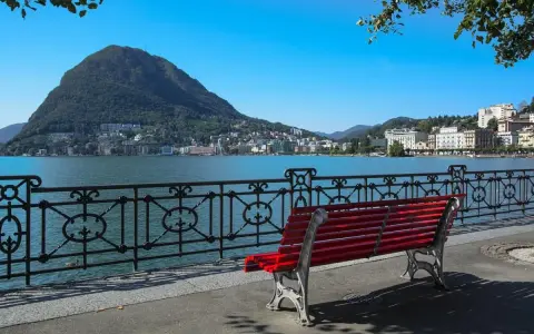 Panoramica del lago di Lugano con Monte San Salvatore sullo sfondo. Una panchina rossa invita a godersi la vista, perfetta per scoprire cosa vedere a Lugano in un giorno.