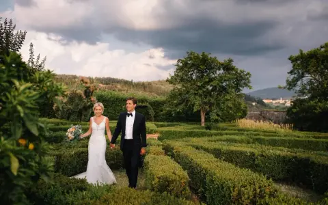 Sposi passeggiano mano nella mano tra i verdi labirinti della **villa di Maiano**, con un bouquet fiorito e uno sfondo di colline toscane.
