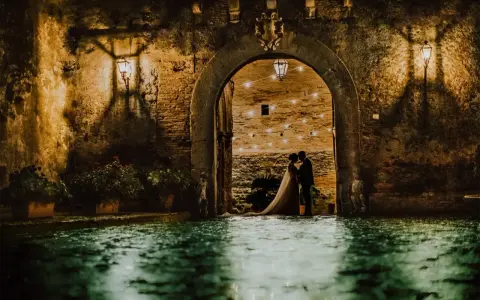 Una coppia di sposi si abbraccia sotto un arco del castello di San Basilio, illuminato da lanterne e luci soffuse. L'acqua riflette l'atmosfera magica.