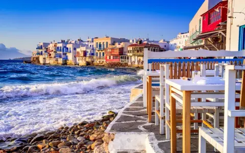 Case vacanza a Mykonos, un'idea per dove andare a vivere all'estero con pochi soldi. Case bianche sul mare, onde che si infrangono sugli scogli, tavoli di un ristorante.