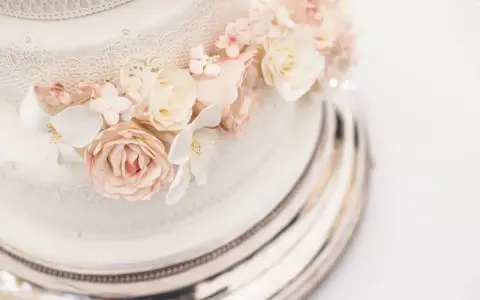 Dettaglio di una torta nuziale a più piani, decorata con pizzo e fiori di zucchero rosa e bianchi, su un elegante vassoio argentato.