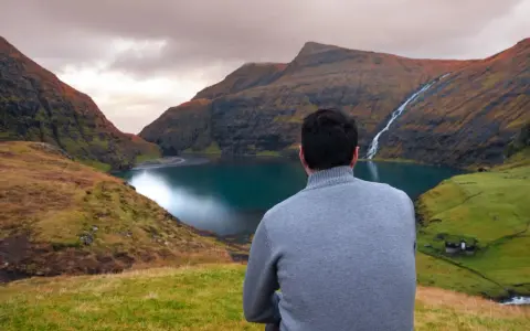 Uomo con maglione grigio ammira un paesaggio mozzafiato delle Isole Faroe, con un lago turchese e una cascata.