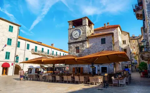 Piazza nel kotor centro storico con torre dell'orologio e tavoli all'aperto di un ristorante.