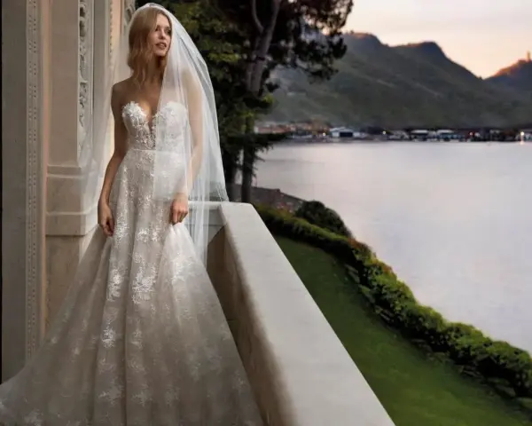 Sposa con abito da sogno, con vari tipi di pizzo che scendono a cascata, su un balcone con vista lago al tramonto.