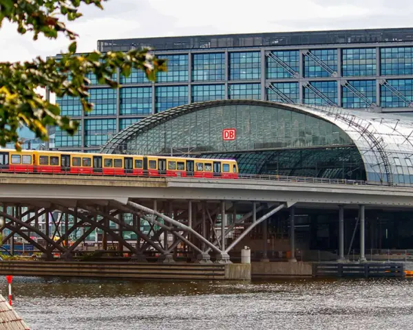 Treno giallo e rosso attraversa un ponte sopra un fiume, con la moderna stazione ferroviaria di Berlino sullo sfondo. Un ottimo modo per capire come muoversi a Berlino.