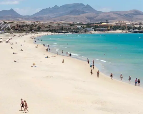 Spiaggia affollata con persone che passeggiano e si rilassano, hotel e montagne sullo sfondo. Questo è il paesaggio di Fuerteventura, dove si trova questo paradiso.