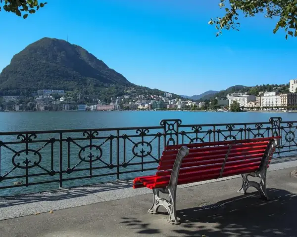 Panoramica del lago di Lugano con Monte San Salvatore sullo sfondo. Una panchina rossa invita a godersi la vista, perfetta per scoprire cosa vedere a Lugano in un giorno.