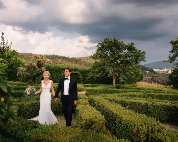 Sposi passeggiano mano nella mano tra i verdi labirinti della **villa di Maiano**, con un bouquet fiorito e uno sfondo di colline toscane.