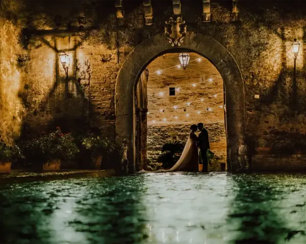 Una coppia di sposi si abbraccia sotto un arco del castello di San Basilio, illuminato da lanterne e luci soffuse. L'acqua riflette l'atmosfera magica.