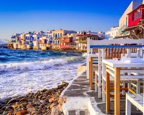 Case vacanza a Mykonos, un'idea per dove andare a vivere all'estero con pochi soldi. Case bianche sul mare, onde che si infrangono sugli scogli, tavoli di un ristorante.