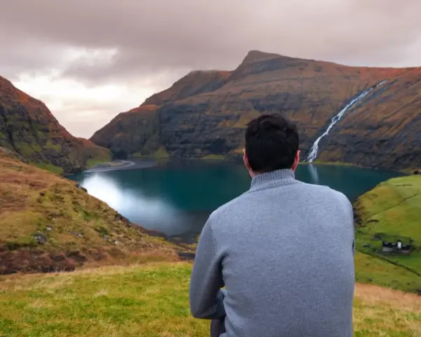 Uomo con maglione grigio ammira un paesaggio mozzafiato delle Isole Faroe, con un lago turchese e una cascata.
