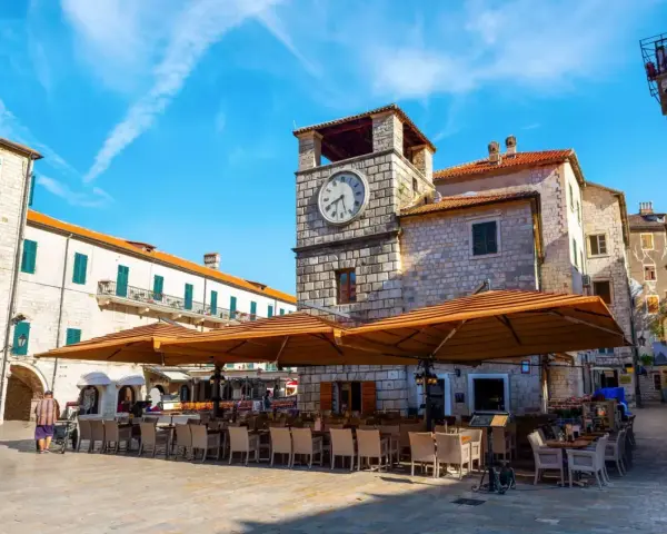 Piazza nel kotor centro storico con torre dell'orologio e tavoli all'aperto di un ristorante.