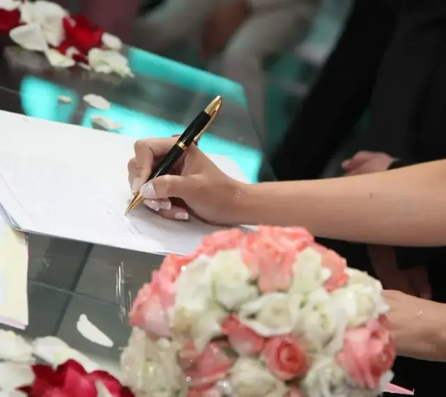 Mano di una sposa che firma un documento, forse un accordo di separazione dei beni, con un bouquet di rose in primo piano.