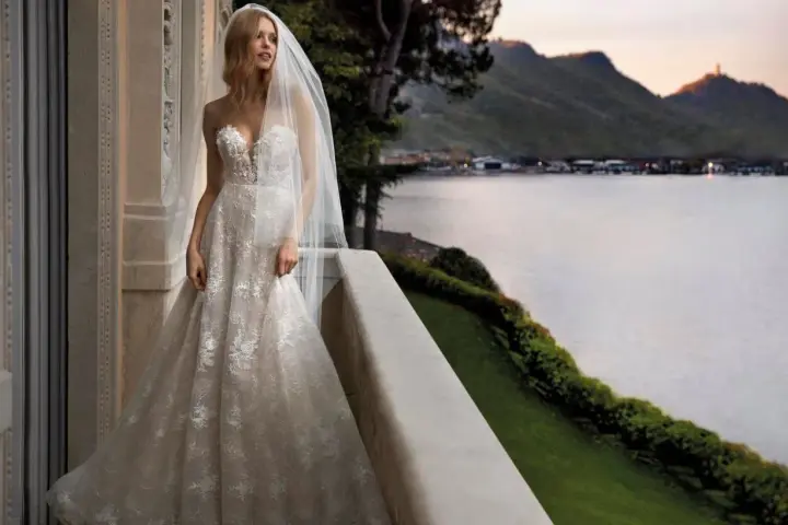 Sposa con abito da sogno, con vari tipi di pizzo che scendono a cascata, su un balcone con vista lago al tramonto.